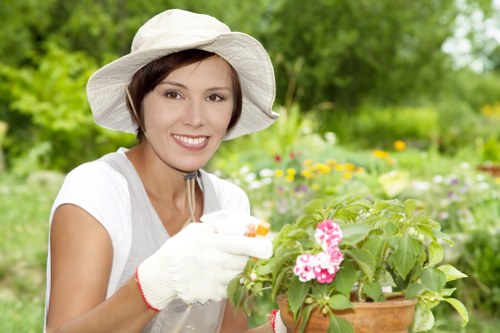 A woman wearing a wide-brimmed white gardening hat, white gloves, and a light-colored sleeveless top is shown tending to a potted flowering plant, likely geraniums, in an outdoor garden setting. The garden features a well-maintained lawn with lush, densely grassed areas in the foreground, bordered by flower beds and shrubbery. Behind her, there are mature green trees and various plants, creating a natural backdrop with vibrant green foliage. The scene is illuminated by natural daylight, suggesting a mild weather day. The garden environment appears tidy and cultivated, with a mix of lawn, flower pots, and natural plant growth, characteristic of a typical residential outdoor space in Rotherhithe or nearby areas. This image reflects professional gardening and maintenance activities that a landscaping service like Lawn Mowing Rotherhithe might undertake, emphasizing the care given to both lawn and garden plants in a domestic setting.
