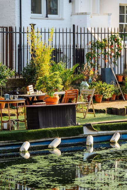 A view of a small city garden featuring a narrow canal with floating buoys and water lilies in the foreground. Behind the canal, there is a paved patio area with outdoor furniture, including a wooden 